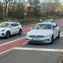 Drei Testfahrzeuge mit Sensoren und Kameras fahren auf einer Straße mit roten Radwegen. Im Vordergrund befindet sich ein weißer VW Passat mit dem Kennzeichen “KL TU 43”, gefolgt von einem weißen Kia Niro mit “Modis”-Branding und einem silbernen Audi. Die Umgebung besteht aus Bäumen, Gebäuden und einer Fußgängerbrücke.