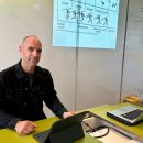 An einem Lehrerpult sitzt ein Lehrer. Rechts neben ihm an der Tafel ist eine Abbildung von einem Speerwurf. Vor ihm auf dem Pult steht sein Tablet.