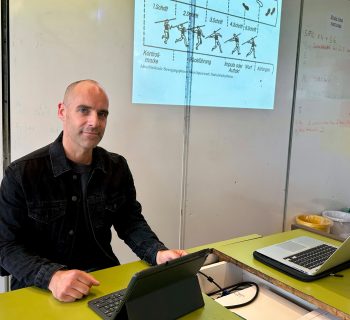 An einem Lehrerpult sitzt ein Lehrer. Rechts neben ihm an der Tafel ist eine Abbildung von einem Speerwurf. Vor ihm auf dem Pult steht sein Tablet.