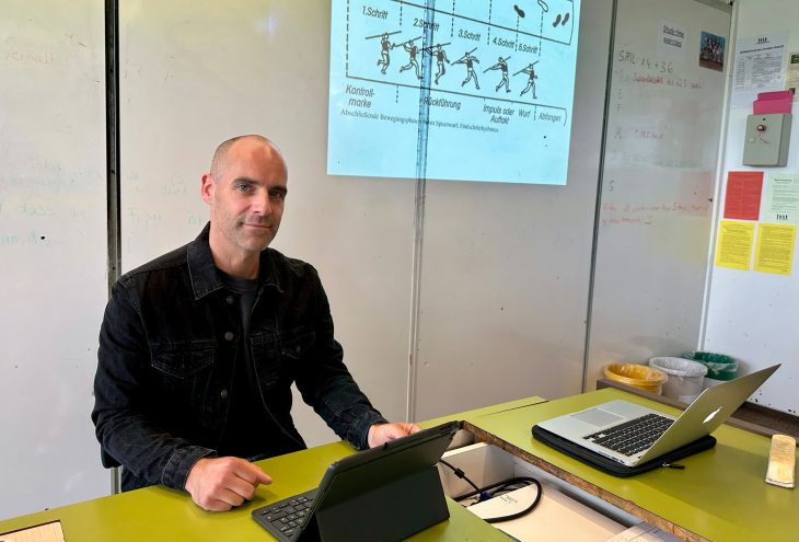 An einem Lehrerpult sitzt ein Lehrer. Rechts neben ihm an der Tafel ist eine Abbildung von einem Speerwurf. Vor ihm auf dem Pult steht sein Tablet.