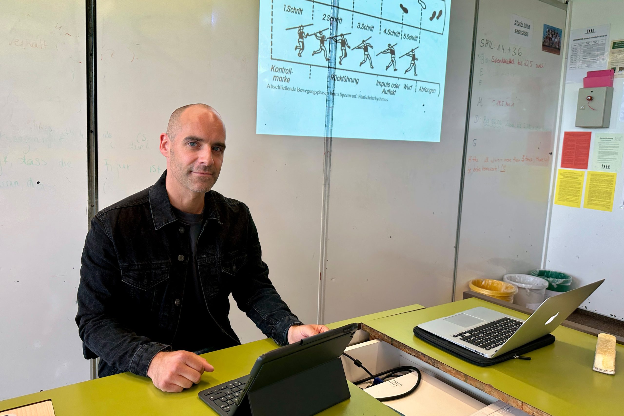 An einem Lehrerpult sitzt ein Lehrer. Rechts neben ihm an der Tafel ist eine Abbildung von einem Speerwurf. Vor ihm auf dem Pult steht sein Tablet.