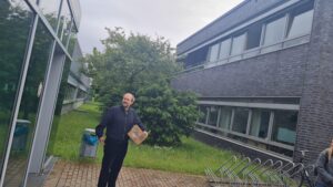 Oskar Schnappauf steht mit einem Paket in der Hand zwischen zwei Hochschulgebäuden. Im Hintergrund sind Fenster, ein Baum, Gras und Fahrradständer zu sehen.