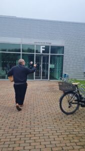 Oskar Schnappauf steht mit einem Paket vor dem Eingang der Biologielabore und zeigt in Richtung Gebäude. Neben dem Eingang sind Fenster, Fahrradständer und ein abgestelltes Fahrrad zu sehen.