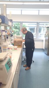 Oskar Schnappauf steht in einem Labor zwischen Arbeitsflächen, Geräten und Regalen und packt ein Paket aus. Im Raum sind Laborutensilien, Flaschen und Arbeitsplätze zu sehen.
