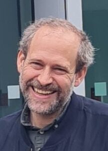 Nahaufnahme von Prof. Dr. Oskar Schnappauf. Er blickt lächelnd in die Kamera, trägt eine dunkle Jacke und steht vor einer Glasfassade.