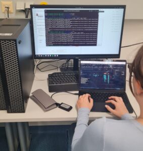 Eine studentische Hilfskraft sitzt an einem Arbeitsplatz im Labor vor Laptop und großem Monitor. Auf den Bildschirmen sind Code, Dateien und wissenschaftliche Arbeitsoberflächen zu sehen.